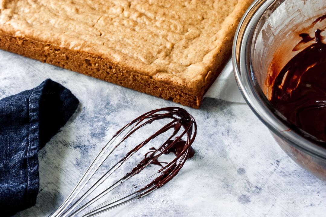 whisk with melted chocolate on it with baked salted caramel blondies in the background