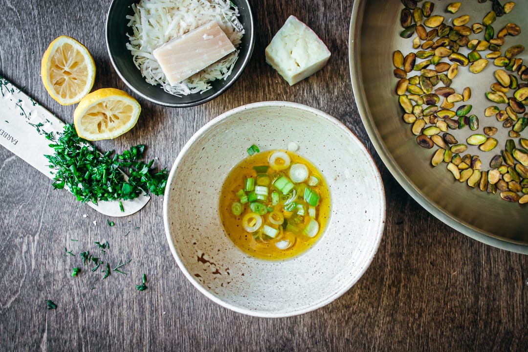 green onions and olive oil mixed in a bowl surrounded by cheese, lemons, herbs, and pistachios