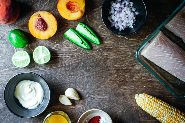 Ingredients for baked fish tacos with peach, corn, and jalapeño salsa arranged in a circle on a wood background