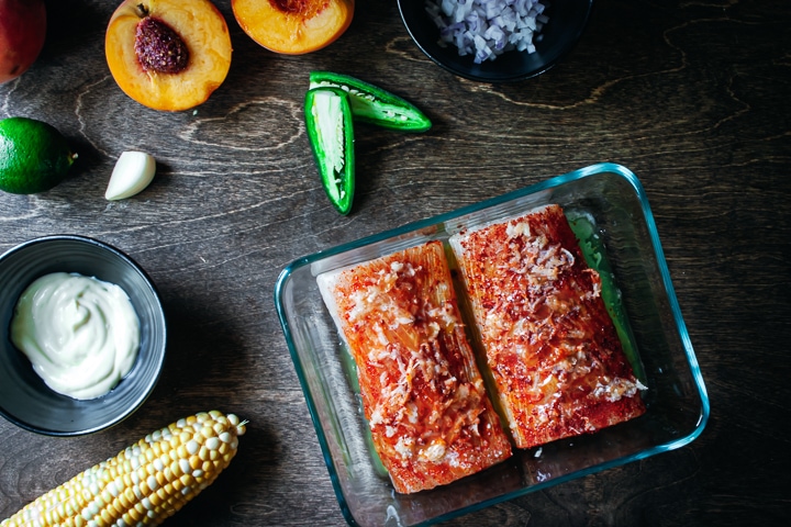 seasoned fish in a glass baking dish with peaches, jalapeños, red onion, and corn on the side