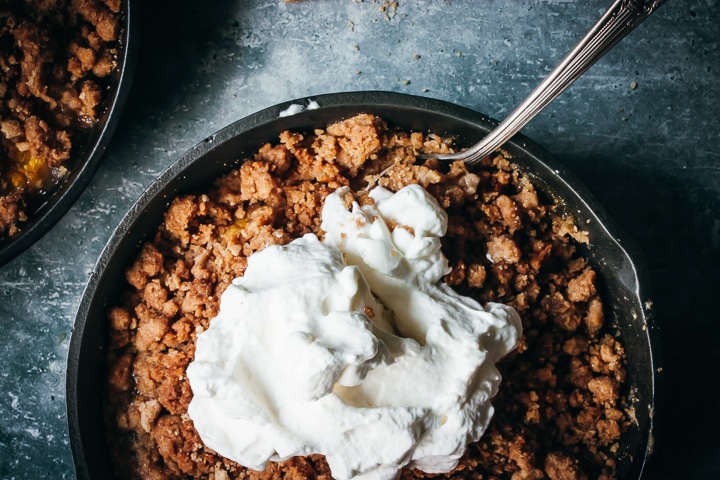 peach crumble for two in a cast iron skillet topped with whipped cream and two spoons on the side