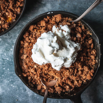 peach crumble for two in a cast iron skillet topped with whipped cream and two spoons