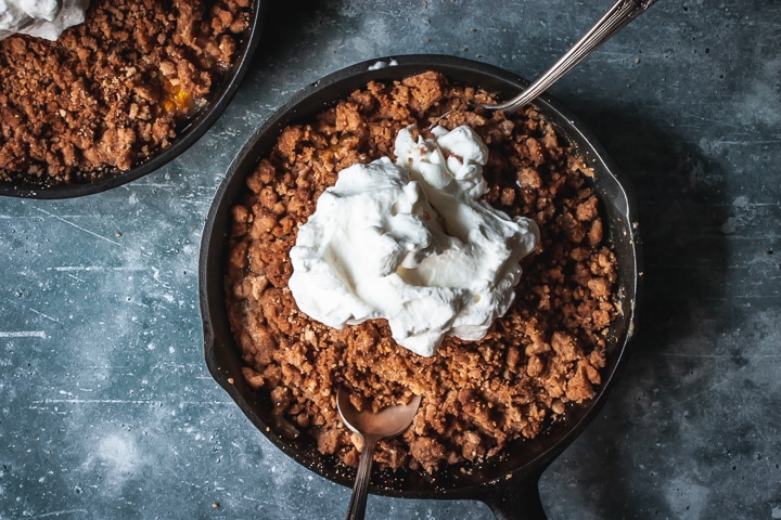 peach crumble for two in a cast iron skillet topped with whipped cream and two spoons