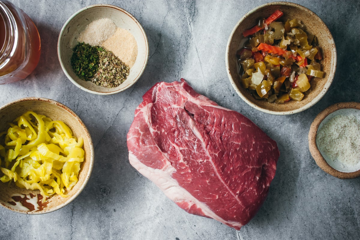 ingredients for Italian beef in different size bowls surrounding a chuck roast