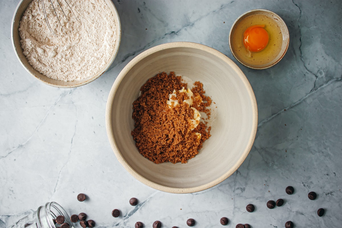 brown sugar and butter in a large bowl surrounded by flour mixture, an egg, and chocolate chips