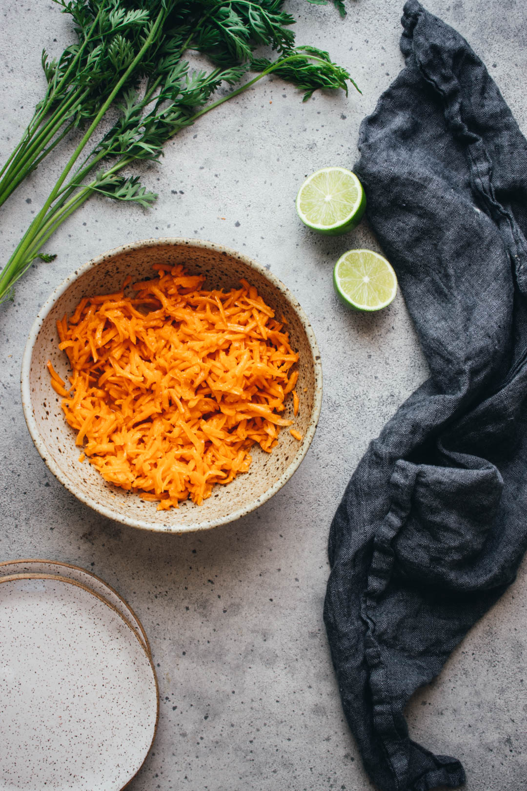 shredded carrot salad in a serving bowl with limes, plates, and a napkin on the side