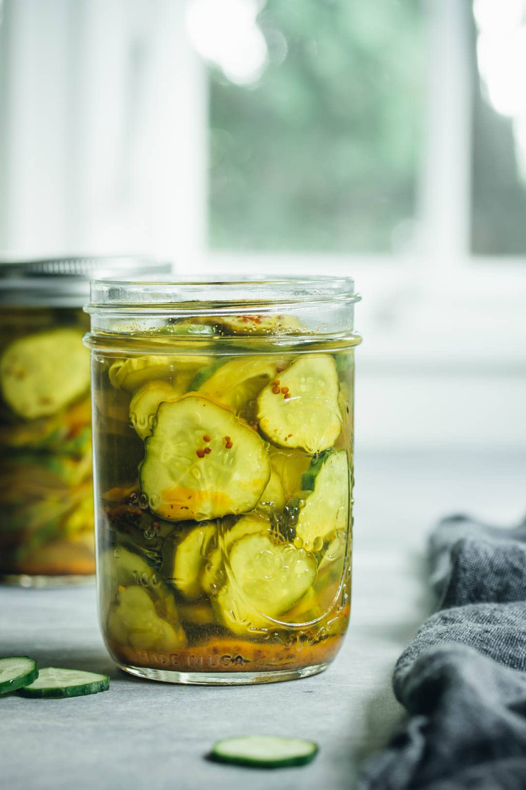 2 jars filled with refrigerator bread and butter pickles with 3 cucumbers and a napkin on side