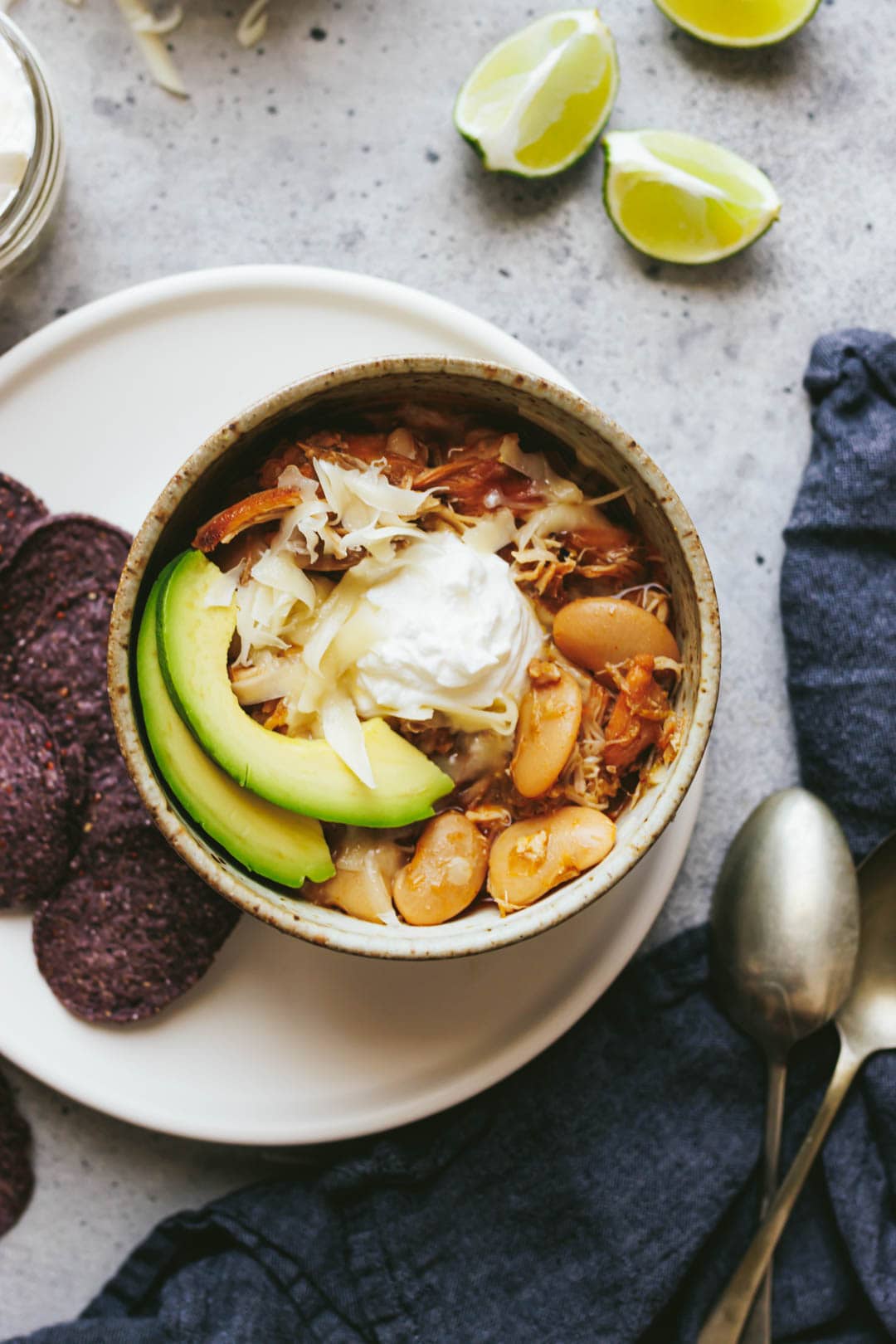 slow cooker white bean chicken chili in a bowl topped with avocado, sour cream, and cheese with tortilla chips and lime wedges on the side