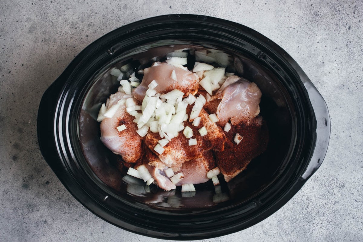 bowl of slow cooker filled with chicken, spices, salt, and chopped onions.